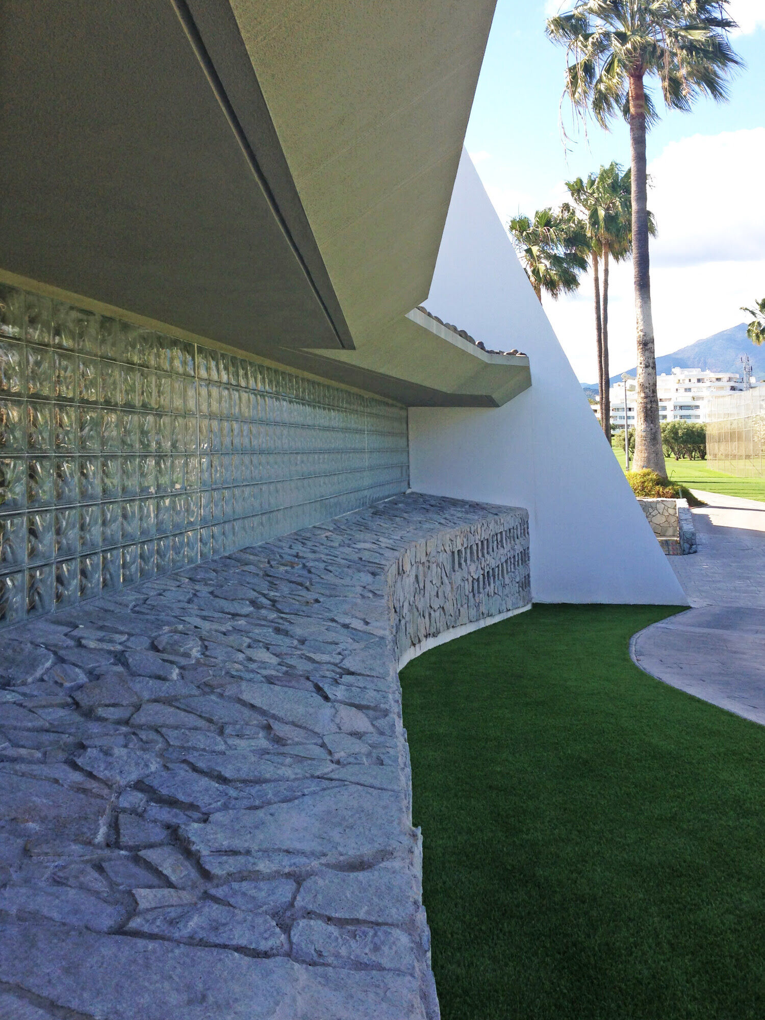 Low-angle detail of the clubhouse exterior wall with textured stone plinth, glass blocks and cantilevered white roof